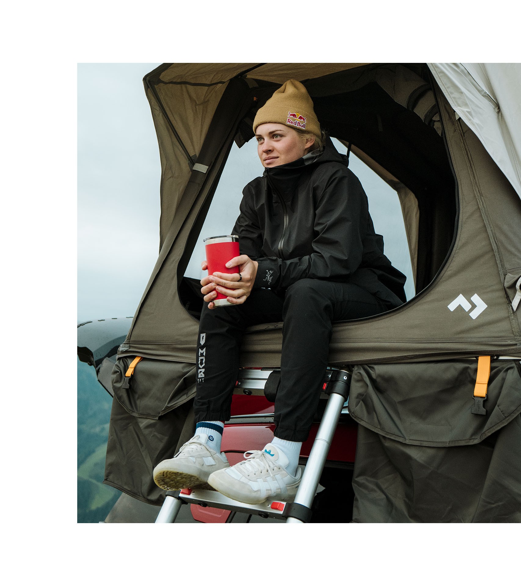 In warm clothing and a Redbull beanie, Vali holds a red cup and sits atop the step ladder leading into a roof tent on the new MINI John Cooper Works Countryman.